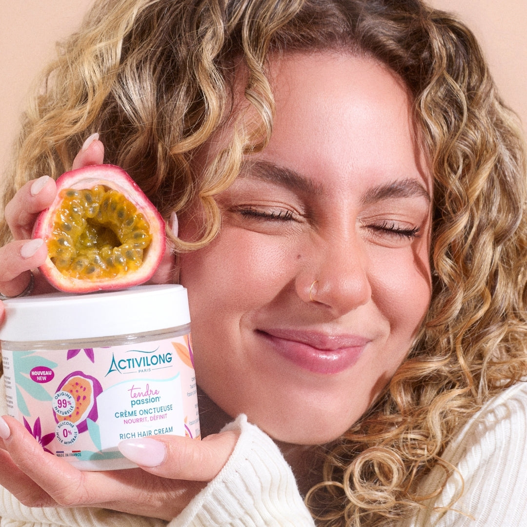 Visuel d'une femme aux cheveux bouclés, yeux clos, tenant le pot Crème onctueuse Tendre Passion et un fruit de la passion coupé près du visage