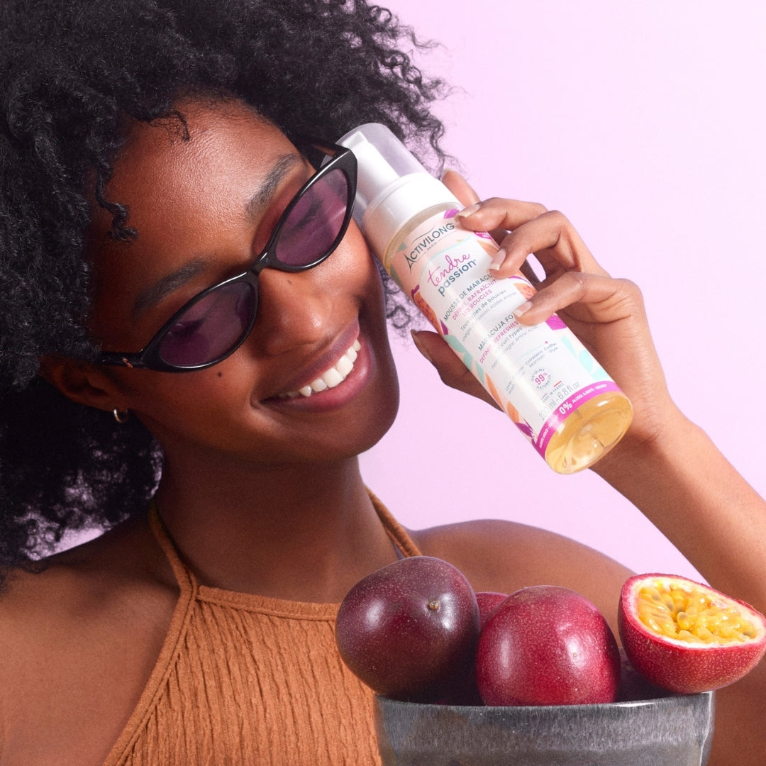 Femme souriante aux cheveux bouclés et lunettes de soleil présentant le flacon-pousse-mousse de Mousse de maracuja Tendre Passion d'Activilong, avec des fruits de la passion entiers et coupé au premier plan, sur fond mauve.