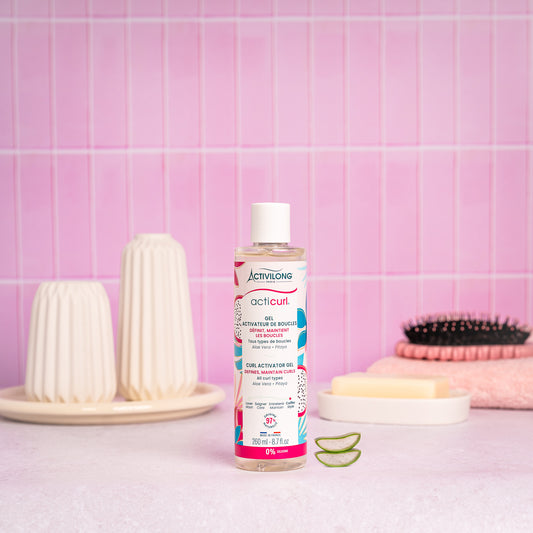 Visuel du flacon Gel activateur de boucles Acticurl Activilong posé sur un plan de salle de bain rose, à côté de tranches d'aloe vera