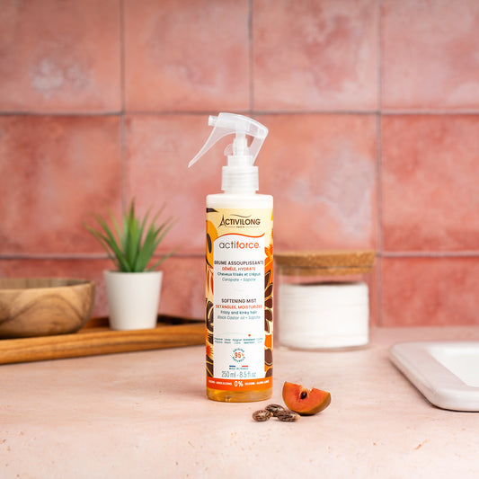 Visuel d'un Flacon de la Brume Assouplissante Actiforce sur un plan de salle de bain terracotta, avec graines de carapate et tranche de sapote en arrière plan