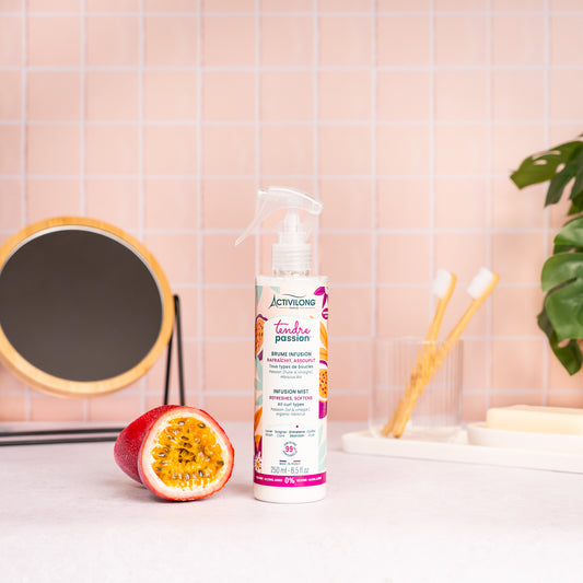 Visuel du spray Brume infusion Tendre Passion posé sur un plan de salle de bain rose, à côté d'un fruit de la passion coupé en deux