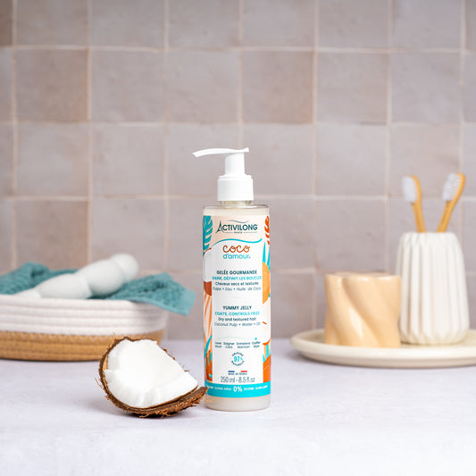 Flacon-pompe de Gelée gourmande Coco d'Amour posé sur un plan de salle de bain, une demi-noix de coco fraîche à côté, avec un panier garni d'une serviette turquoise, un gobelet et des brosses à dents en arrière-plan, sur fond de carrelage beige.