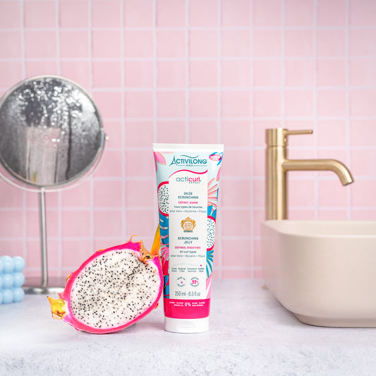 Visuel d'un Tube de la Gelée Scrunching Acticurl Expert sur un plan de salle de bain rose, avec miroir, vasque et demi-pitaya en arrière plan