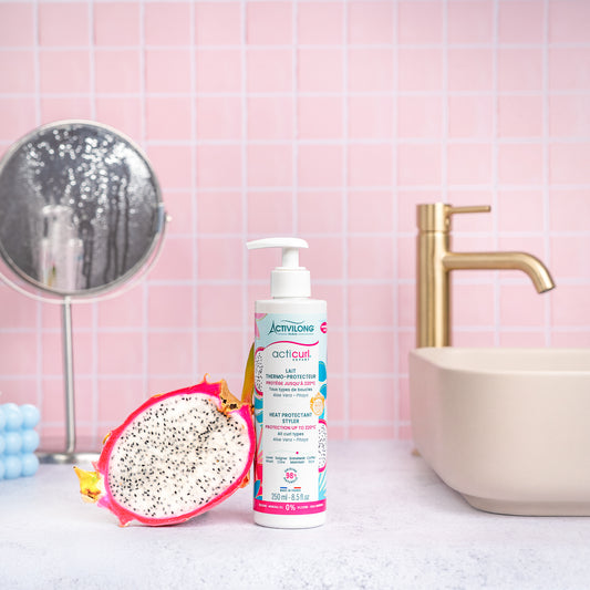 Visuel d'un Flacon du Lait Thermo-Protecteur Acticurl Expert sur un plan de salle de bain rose, avec miroir, vasque et demi-pitaya en arrière plan