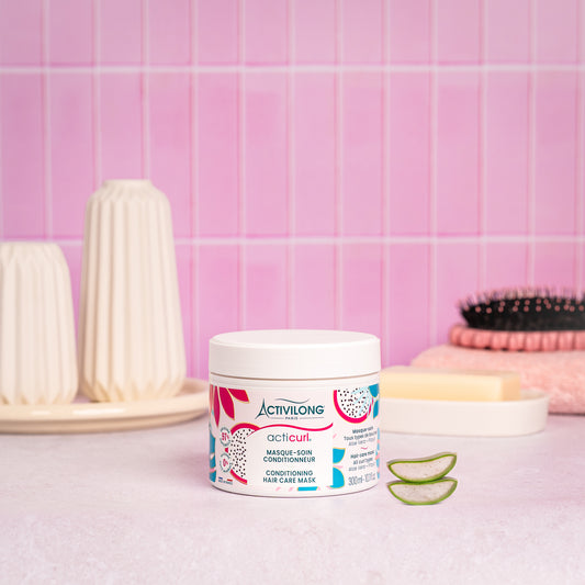 Visuel d'un Pot du Masque-soin Conditionneur Acticurl sur un plan de salle de bain rose, avec brosse, savon et tranches d'aloe vera en arrière plan