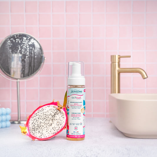 Visuel d'un Flacon de la Mousse Bouclante Acticurl Expert sur un plan de salle de bain rose, avec miroir, vasque et demi-pitaya en arrière plan