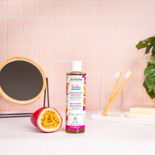 Visuel du flacon Shampooing fruité Tendre Passion posé sur un plan de salle de bain rose, à côté d'un fruit de la passion coupé en deux
