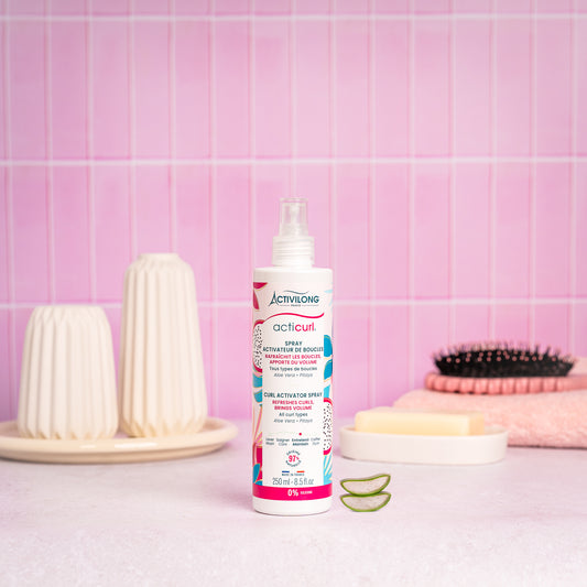 Visuel d'un Flacon du Spray Activateur de Boucles Acticurl sur un plan de salle de bain rose, avec brosse, savon et tranches d'aloe vera en arrière plan