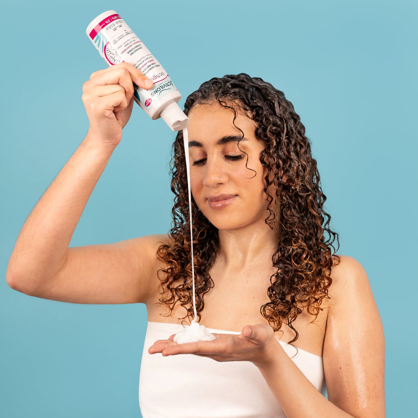 Visuel d'une femme aux cheveux bouclés humides versant l'Après-shampooing sublimateur Acticurl Expert Activilong dans le creux de sa main, sur fond bleu clair