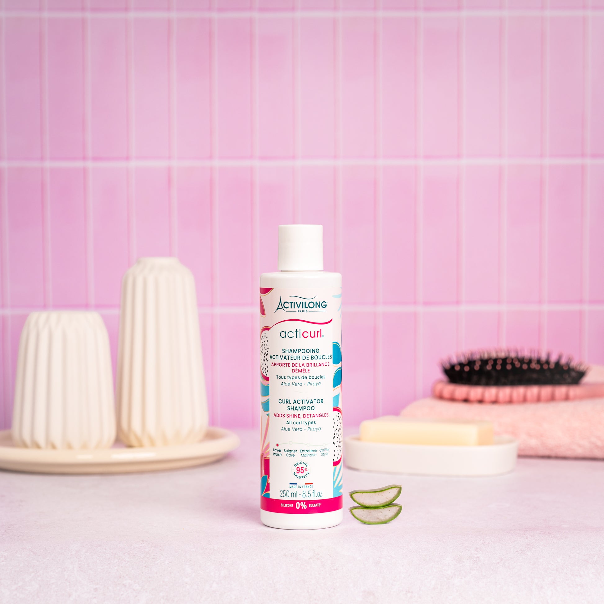 Visuel d'un Flacon du Shampoing Activateur de Boucles Acticurl sur un plan de salle de bain rose, avec brosse, savon et tranches d'aloe vera en arrière plan