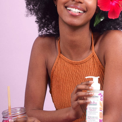 Femme souriante aux cheveux frisés, une fleur d'hibiscus rose dans les cheveux, présentant le flacon-pompe de Gelée fondante Tendre Passion, sur fond mauve.