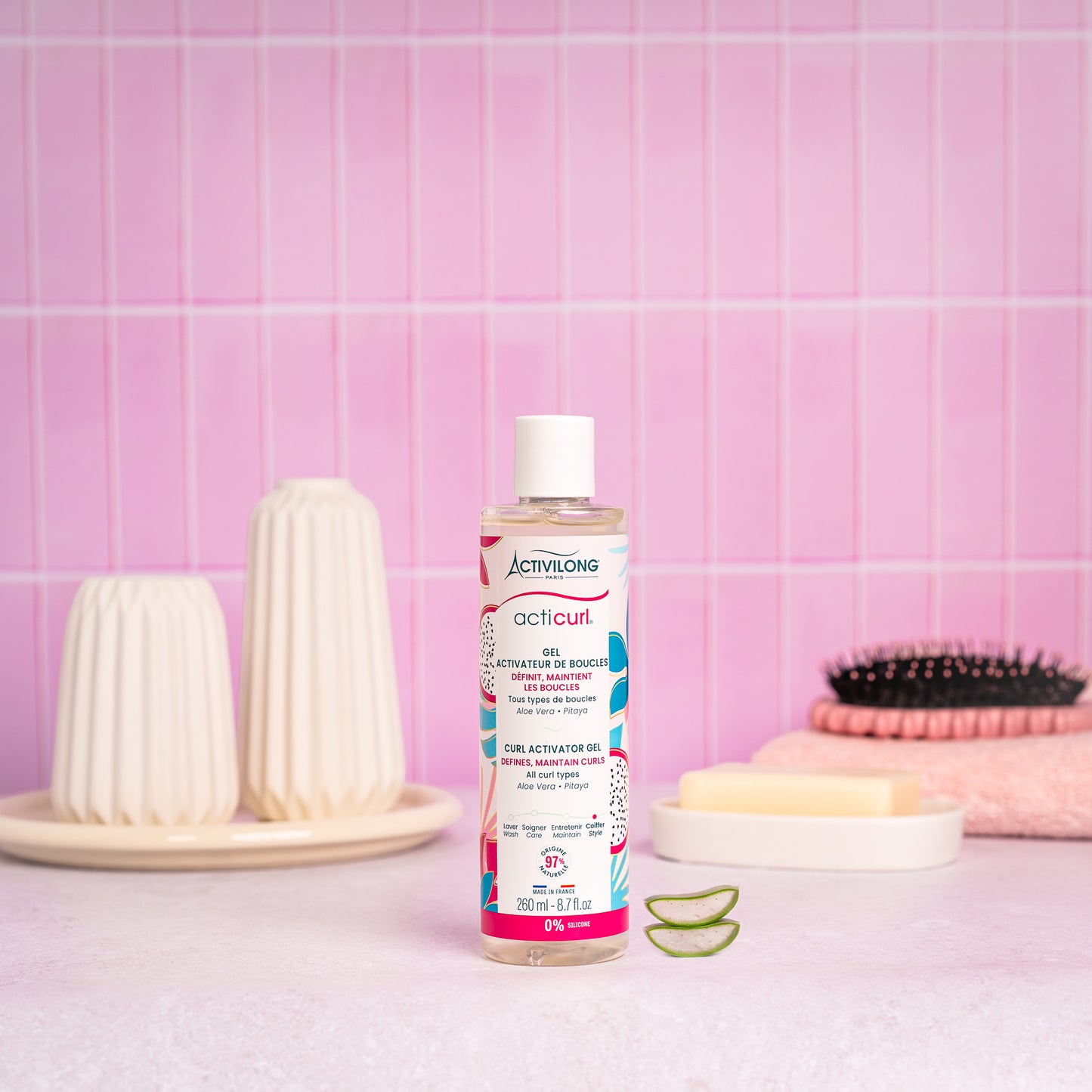 Visuel du flacon Gel activateur de boucles Acticurl Activilong posé sur un plan de salle de bain rose, à côté de tranches d'aloe vera