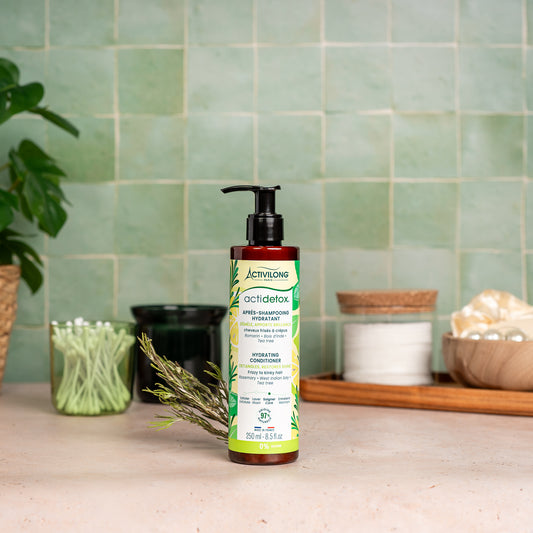 Visuel d'un Flacon de l'Après-Shampooing Hydratant Actidetox sur un plan de salle de bain vert, avec brin de romarin et accessoires en arrière plan