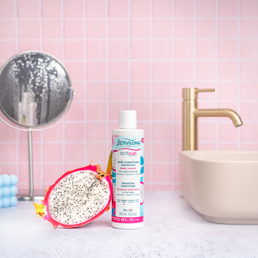 Visuel d'un Flacon de l'Après-Shampooing Sublimateur Acticurl Expert sur un plan de salle de bain rose, avec miroir, vasque et demi-pitaya en arrière plan