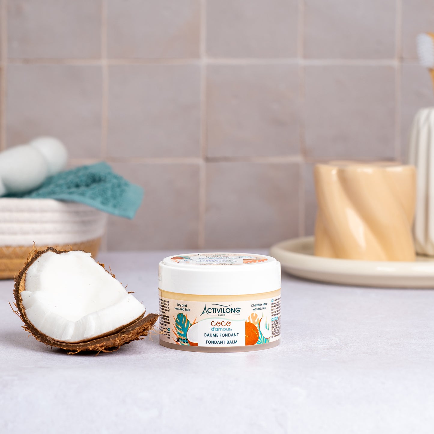 Pot de Baume fondant Coco d'Amour posé sur un plan de salle de bain, une demi-noix de coco fraîche à côté, avec un panier garni d'une serviette turquoise et un gobelet en arrière-plan, sur fond de carrelage beige.