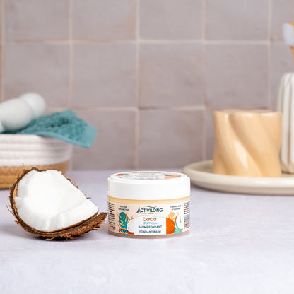Pot de Baume fondant Coco d'Amour posé sur un plan de salle de bain, une demi-noix de coco fraîche à côté, avec un panier garni d'une serviette turquoise et un gobelet en arrière-plan, sur fond de carrelage beige.