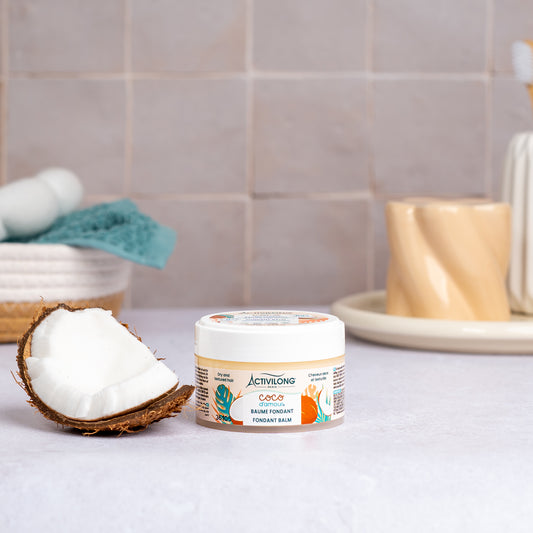 Pot de Baume fondant Coco d'Amour posé sur un plan de salle de bain, une demi-noix de coco fraîche à côté, avec un panier garni d'une serviette turquoise et un gobelet en arrière-plan, sur fond de carrelage beige.