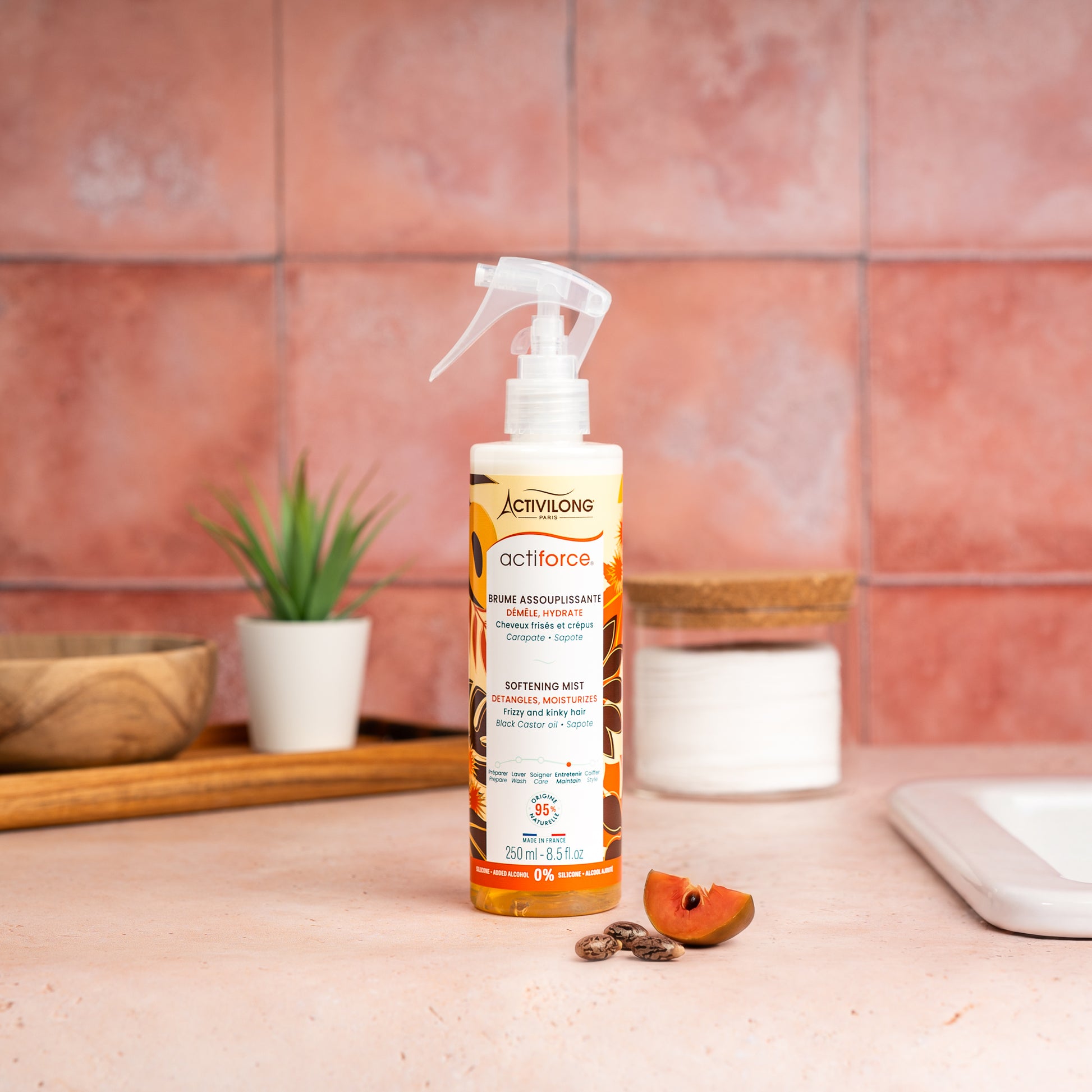 Visuel d'un Flacon de la Brume Assouplissante Actiforce sur un plan de salle de bain terracotta, avec graines de carapate et tranche de sapote en arrière plan