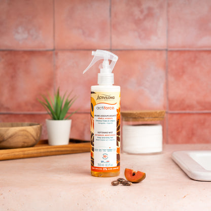 Visuel d'un Flacon de la Brume Assouplissante Actiforce sur un plan de salle de bain terracotta, avec graines de carapate et tranche de sapote en arrière plan