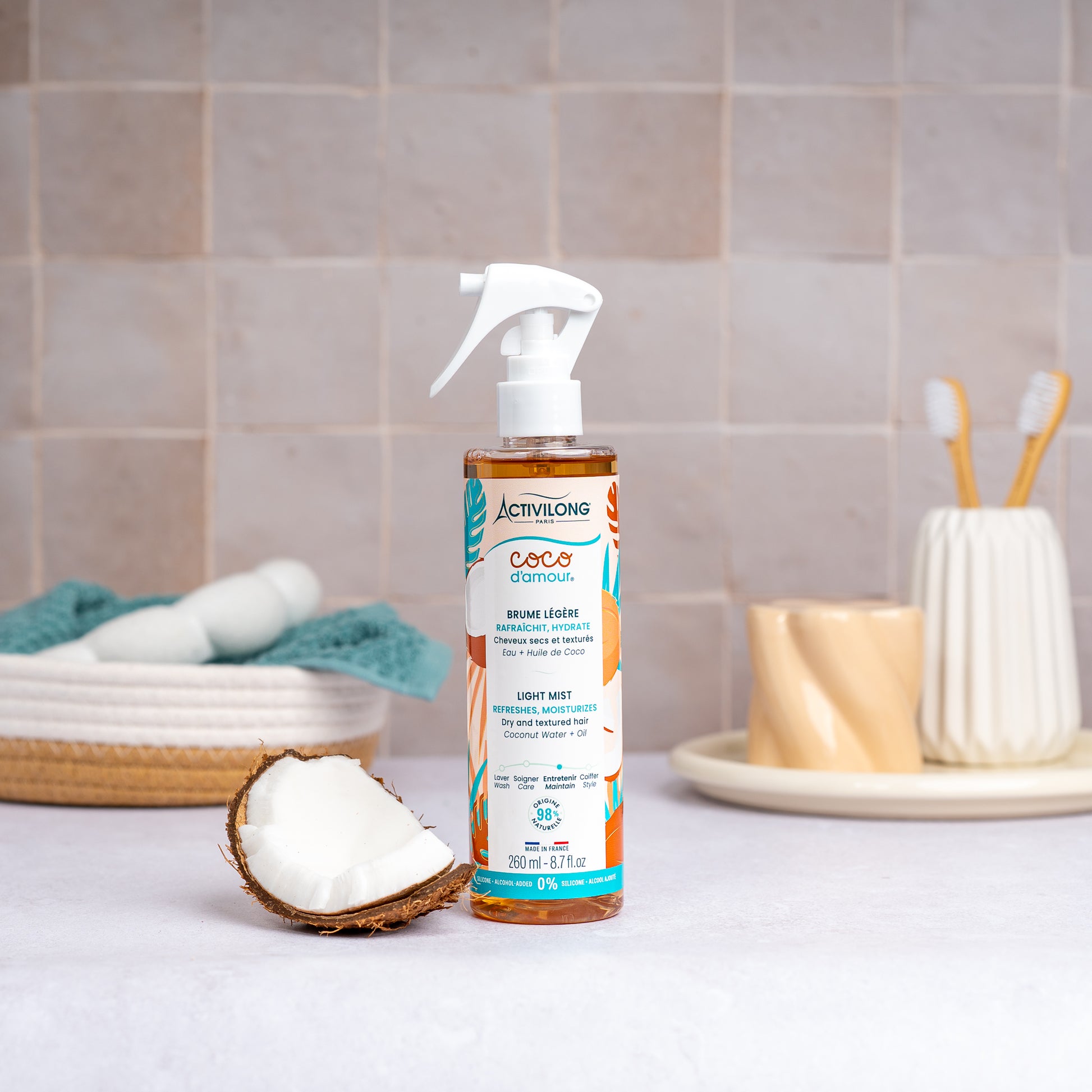 Flacon-spray de Brume légère Coco d'Amour posé sur un plan de salle de bain, une demi-noix de coco fraîche à côté, avec un panier garni d'une serviette turquoise, un gobelet et des brosses à dents en arrière-plan, sur fond de carrelage beige.