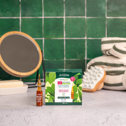 Visuel de la boîte du Complexe Racines Actigro et d'une ampoule, sur un plan de salle de bain vert, avec miroir, brosse de massage et serviettes en arrière plan
