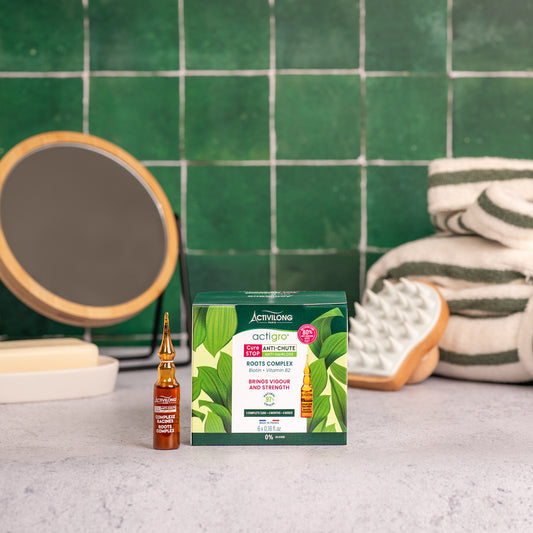 Visuel de la boîte du Complexe Racines Actigro et d'une ampoule, sur un plan de salle de bain vert, avec miroir, brosse de massage et serviettes en arrière plan