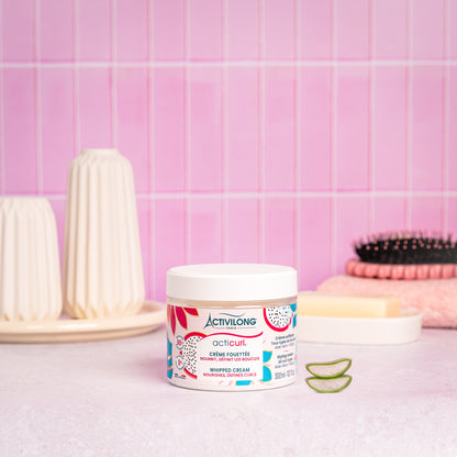 Visuel d’un Pot de la Crème Fouettée Acticurl sur un plan de salle de bain rose, avec brosse, savon et tranches d’aloe vera en arrière plan