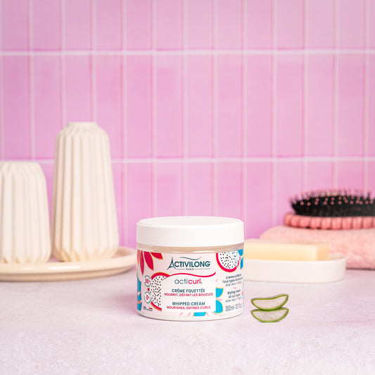 Visuel d’un Pot de la Crème Fouettée Acticurl sur un plan de salle de bain rose, avec brosse, savon et tranches d’aloe vera en arrière plan