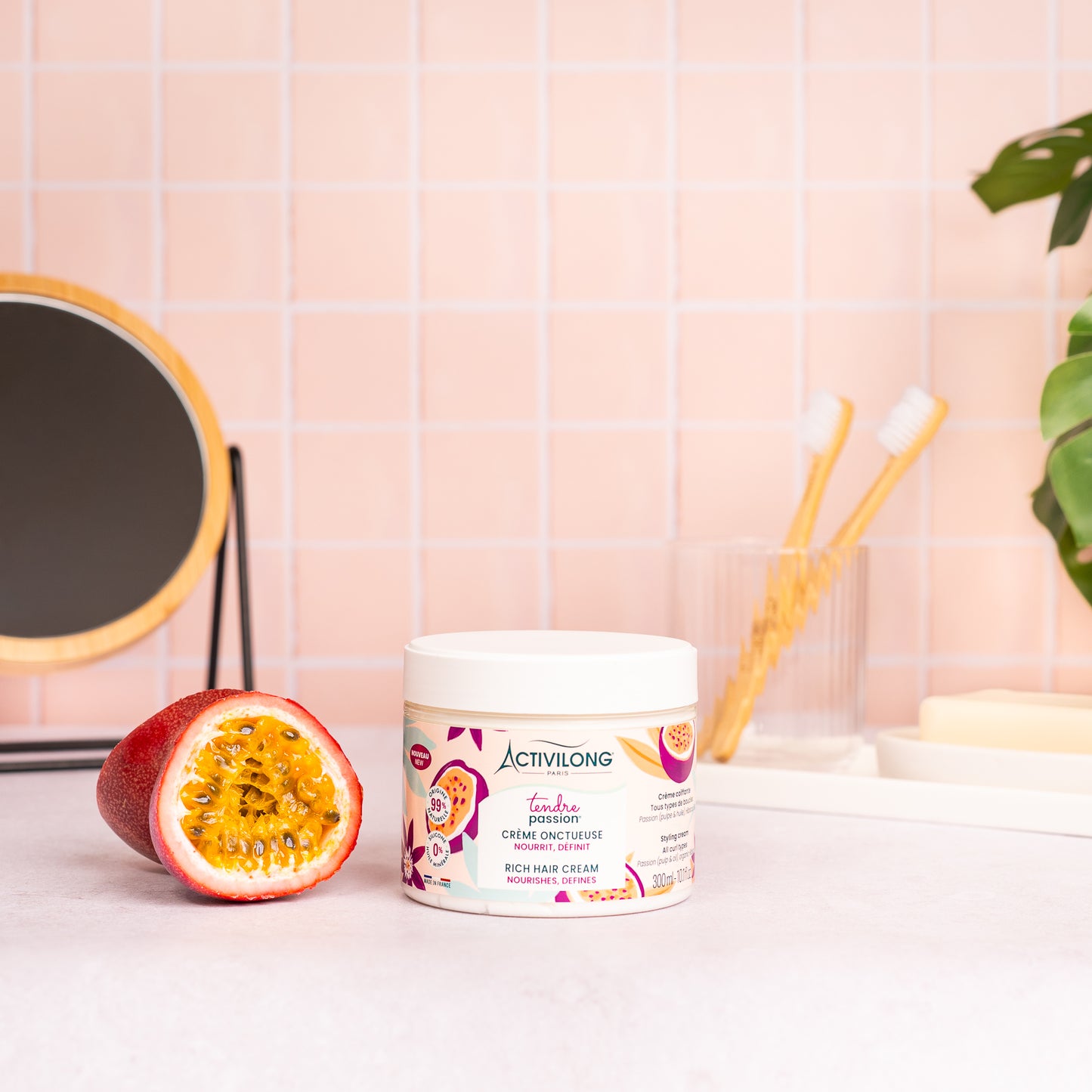 Visuel du pot Crème onctueuse Tendre Passion posé sur un plan de salle de bain rose, à côté d'un fruit de la passion coupé en deux