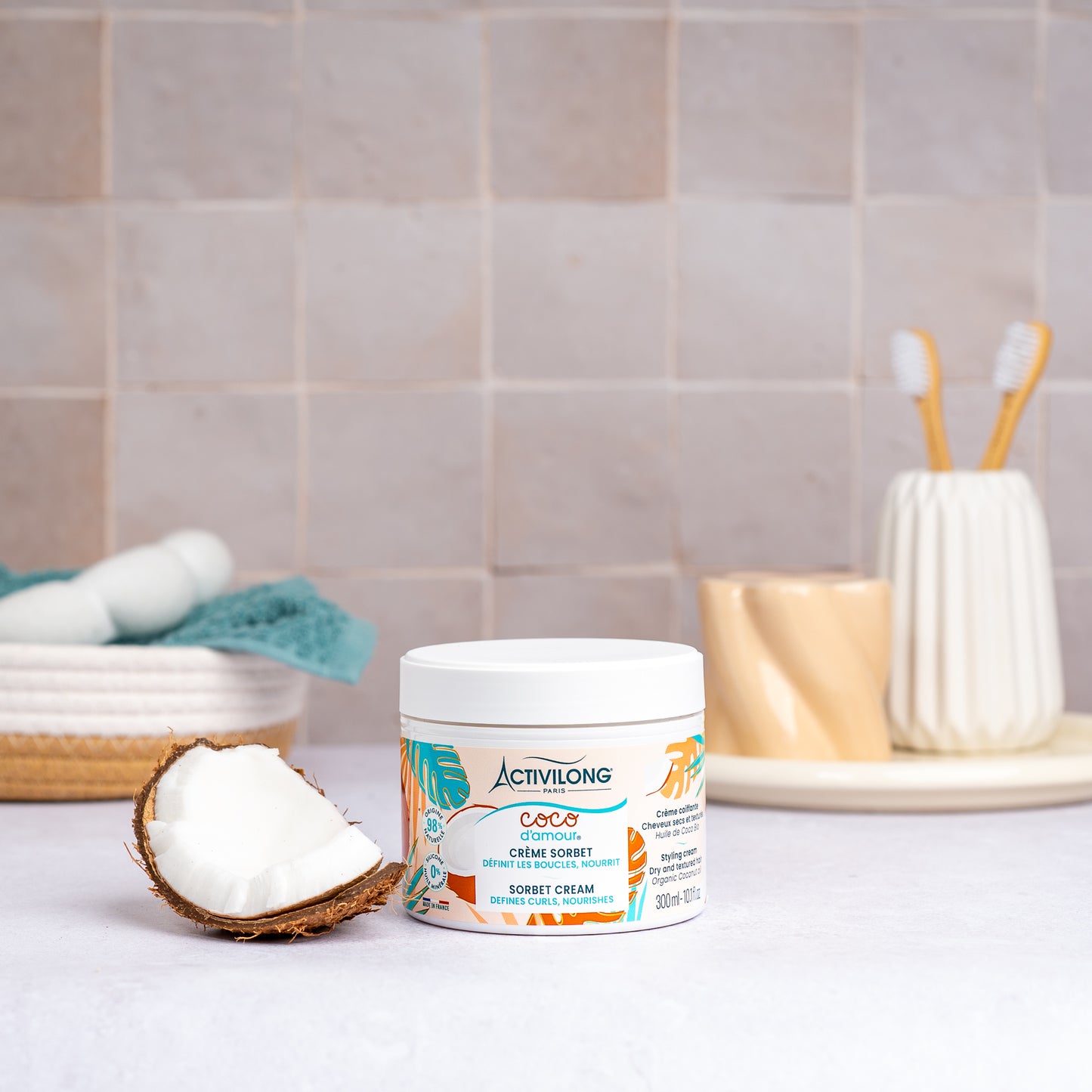 Pot de Crème sorbet Coco d'Amour posé sur un plan de salle de bain, une demi-noix de coco fraîche à côté, avec un panier garni d'une serviette turquoise, un gobelet et des brosses à dents en arrière-plan, sur fond de carrelage beige.