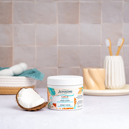 Pot de Crème sorbet Coco d'Amour posé sur un plan de salle de bain, une demi-noix de coco fraîche à côté, avec un panier garni d'une serviette turquoise, un gobelet et des brosses à dents en arrière-plan, sur fond de carrelage beige.
