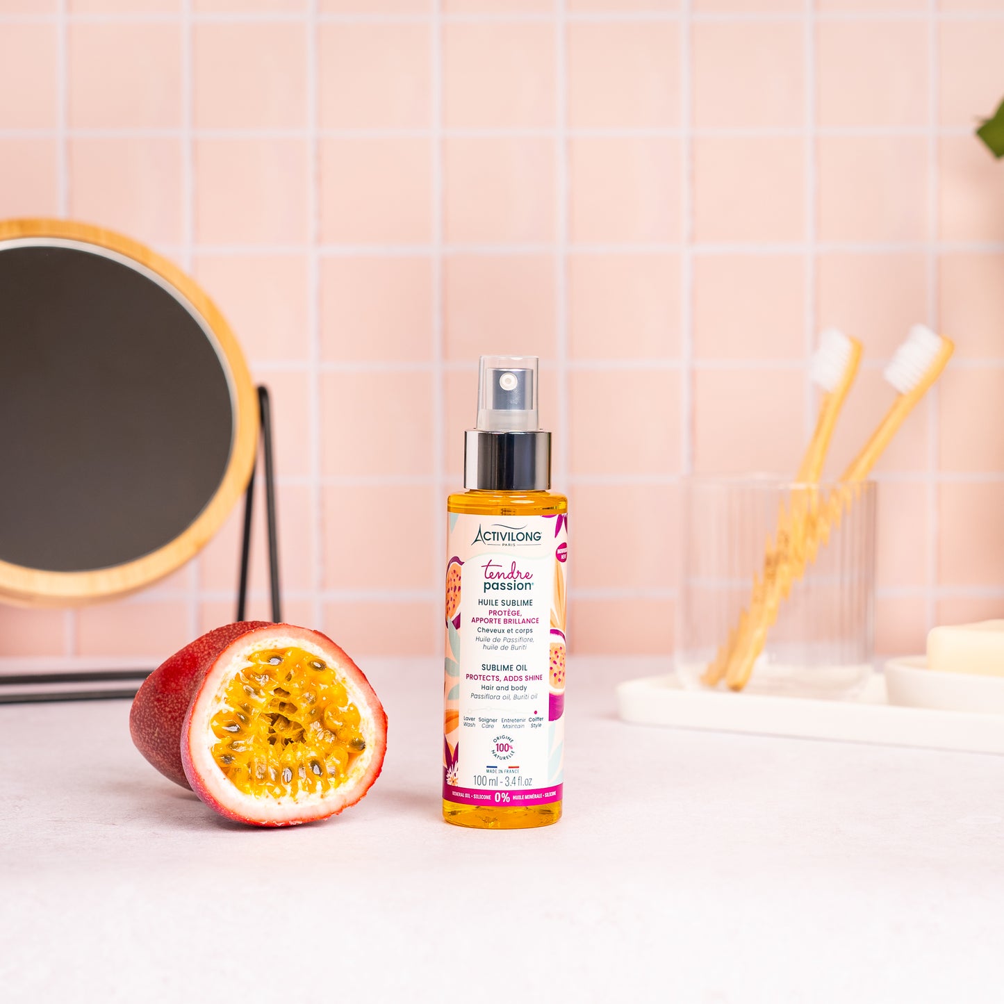 Visuel du spray Huile sublime Tendre Passion posé sur un plan de salle de bain rose, à côté d'un fruit de la passion coupé en deux