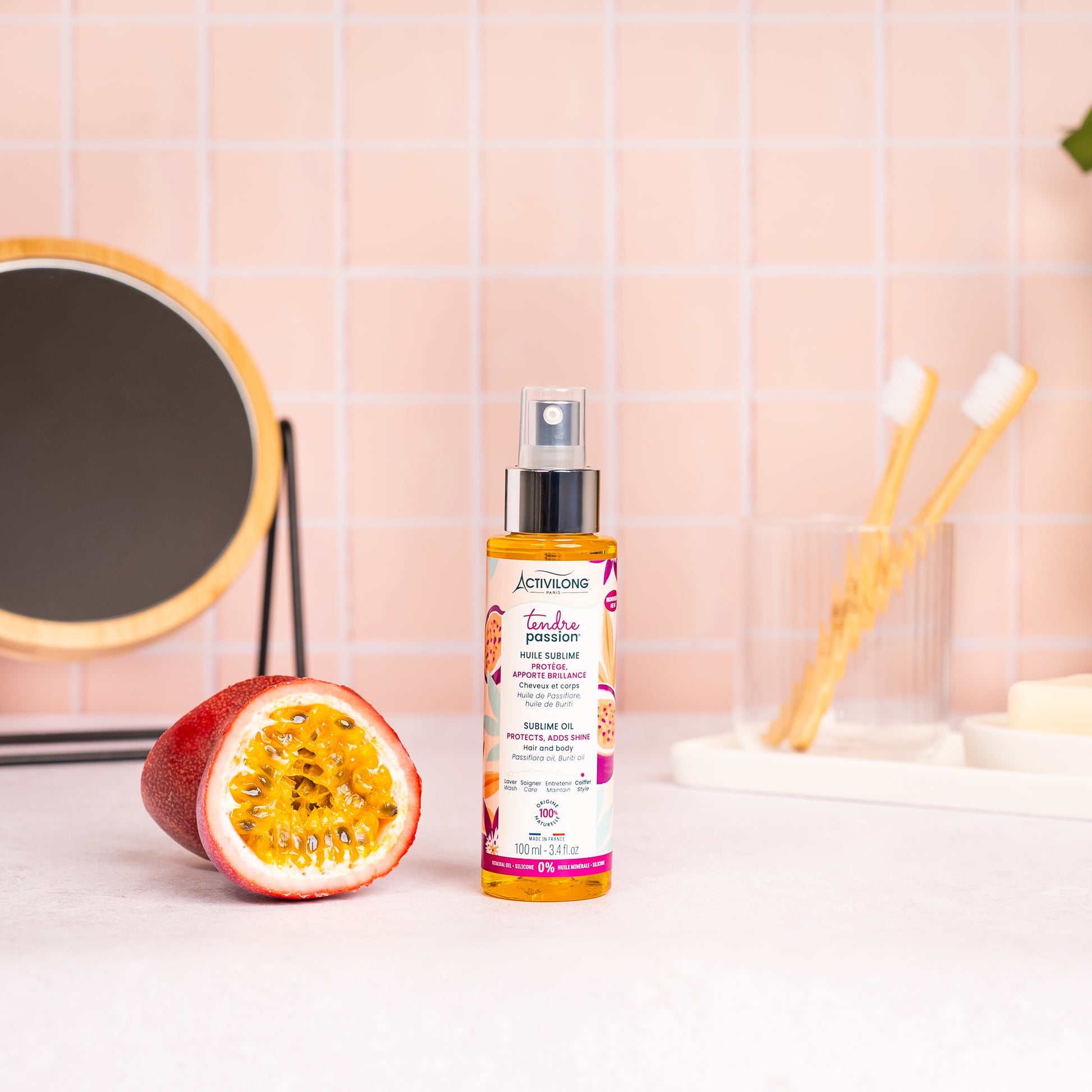 Visuel du spray Huile sublime Tendre Passion posé sur un plan de salle de bain rose, à côté d'un fruit de la passion coupé en deux