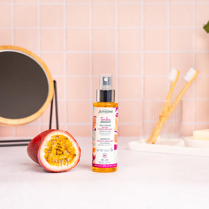 Visuel du spray Huile sublime Tendre Passion posé sur un plan de salle de bain rose, à côté d'un fruit de la passion coupé en deux
