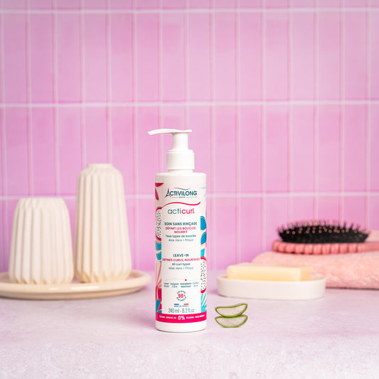Visuel d'un Flacon du Soin Sans Rinçage Acticurl sur un plan de salle de bain rose, avec brosse, savon et tranches d'aloe vera en arrière plan