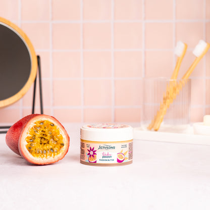 Pot de Passion butter Tendre Passion posé sur un plan de salle de bain, un fruit de la passion coupé révélant sa pulpe à côté, avec un miroir sur pied en bambou, un gobelet et des brosses à dents en arrière-plan, sur fond de carrelage rose poudré.