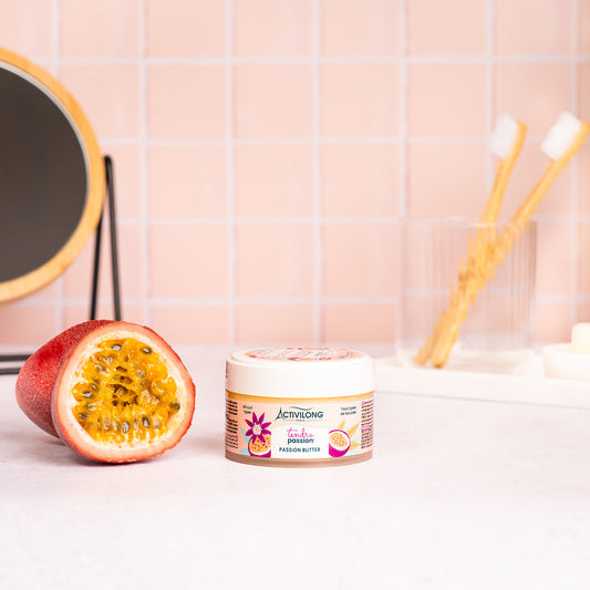 Pot de Passion butter Tendre Passion posé sur un plan de salle de bain, un fruit de la passion coupé révélant sa pulpe à côté, avec un miroir sur pied en bambou, un gobelet et des brosses à dents en arrière-plan, sur fond de carrelage rose poudré.