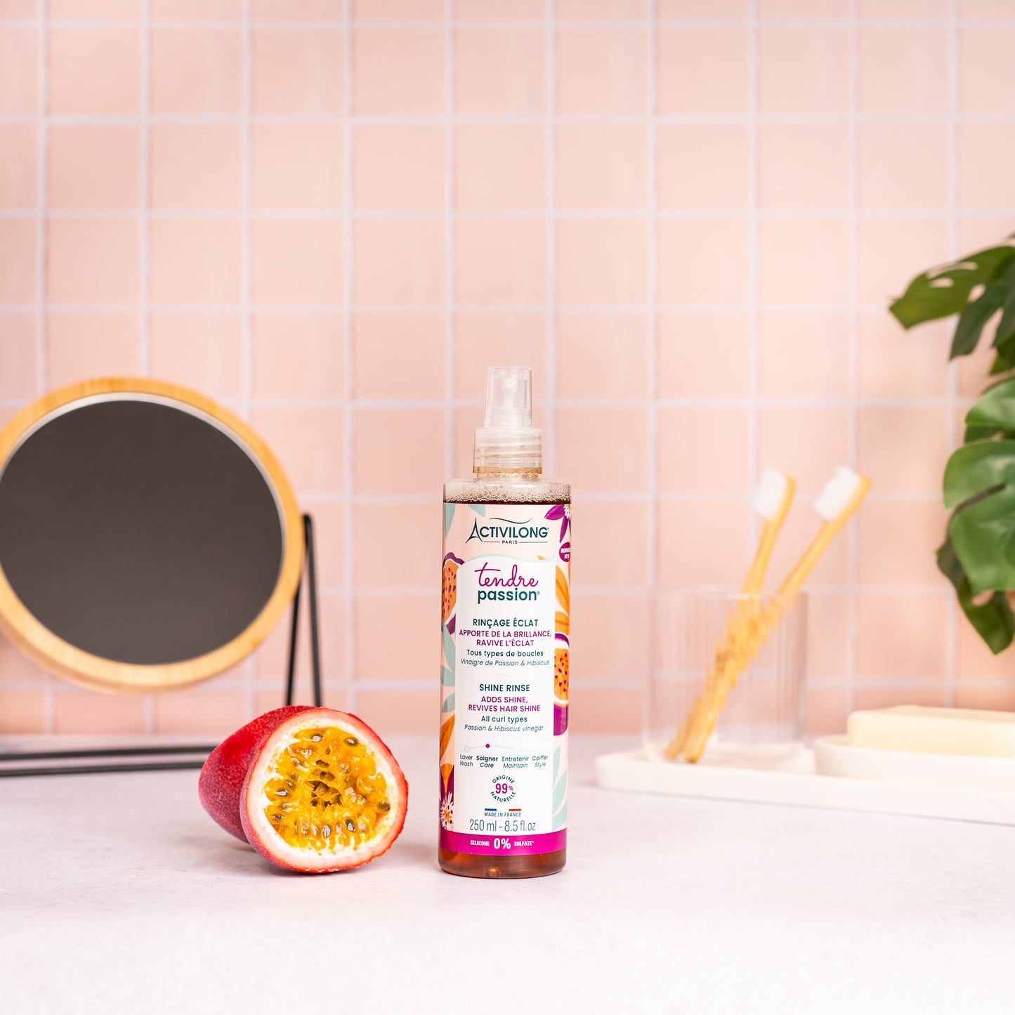 Visuel du spray Rinçage éclat Tendre Passion posé sur un plan de salle de bain rose, à côté d'un fruit de la passion coupé en deux