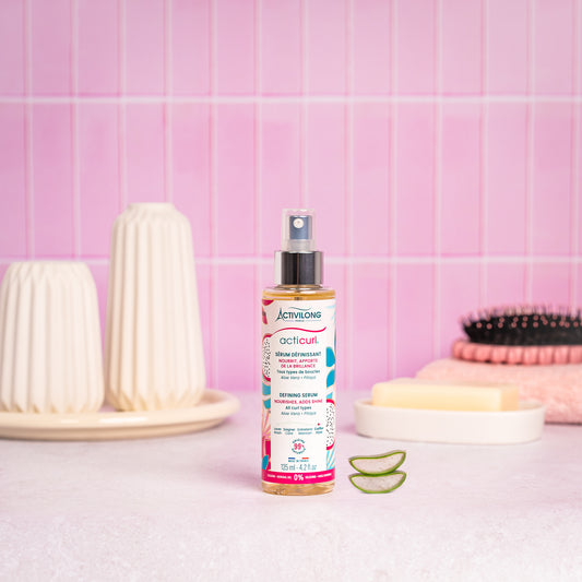 Visuel d'un Flacon du Sérum Définissant Acticurl sur un plan de salle de bain rose, avec brosse, savon et tranches d'aloe vera en arrière plan