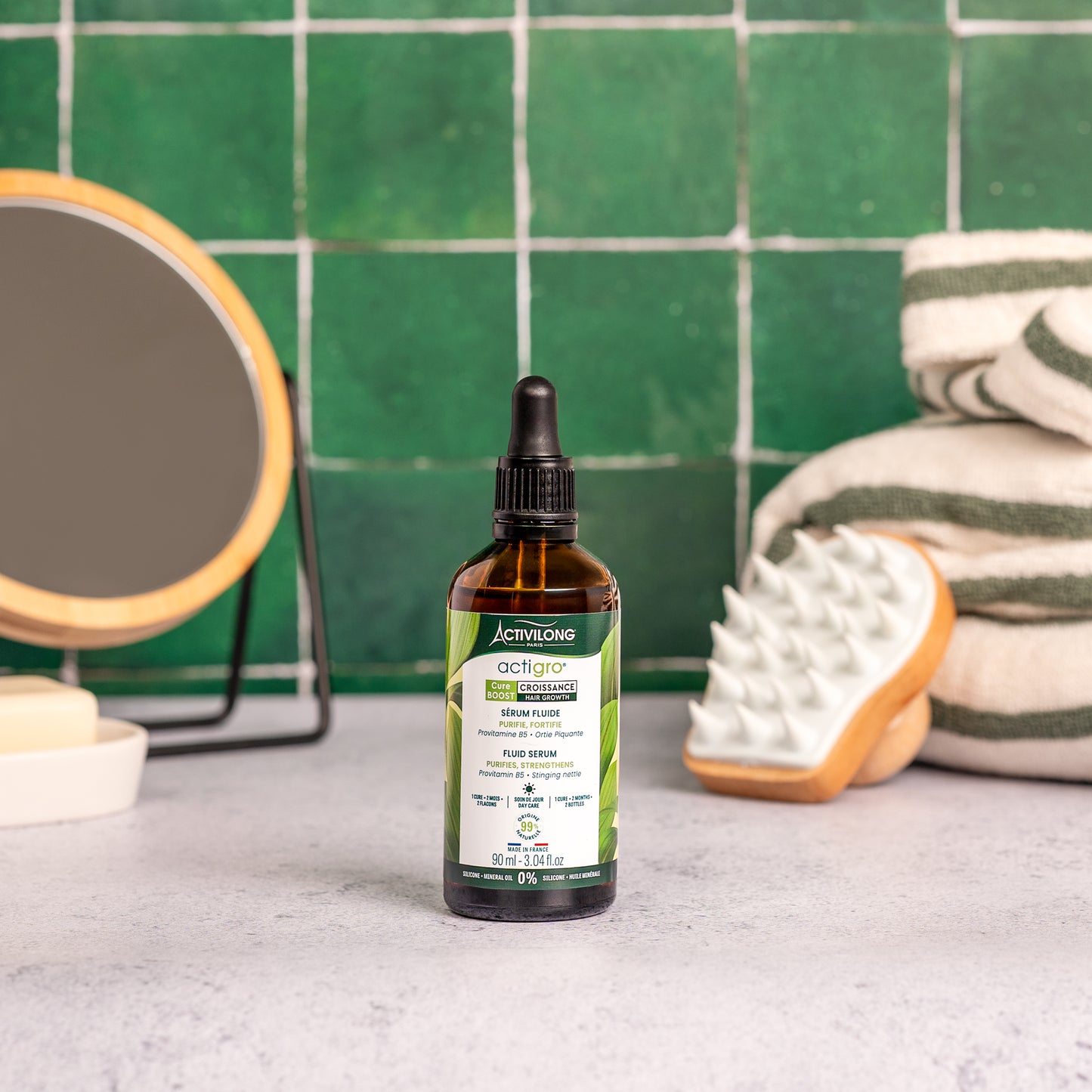Visuel d'un Flacon de 90 ml du Sérum Fluide Actigro sur un plan de salle de bain vert, avec miroir, brosse de massage et serviettes en arrière plan
