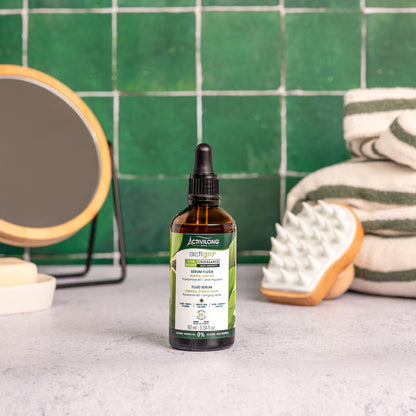 Visuel d'un Flacon de 90 ml du Sérum Fluide Actigro sur un plan de salle de bain vert, avec miroir, brosse de massage et serviettes en arrière plan