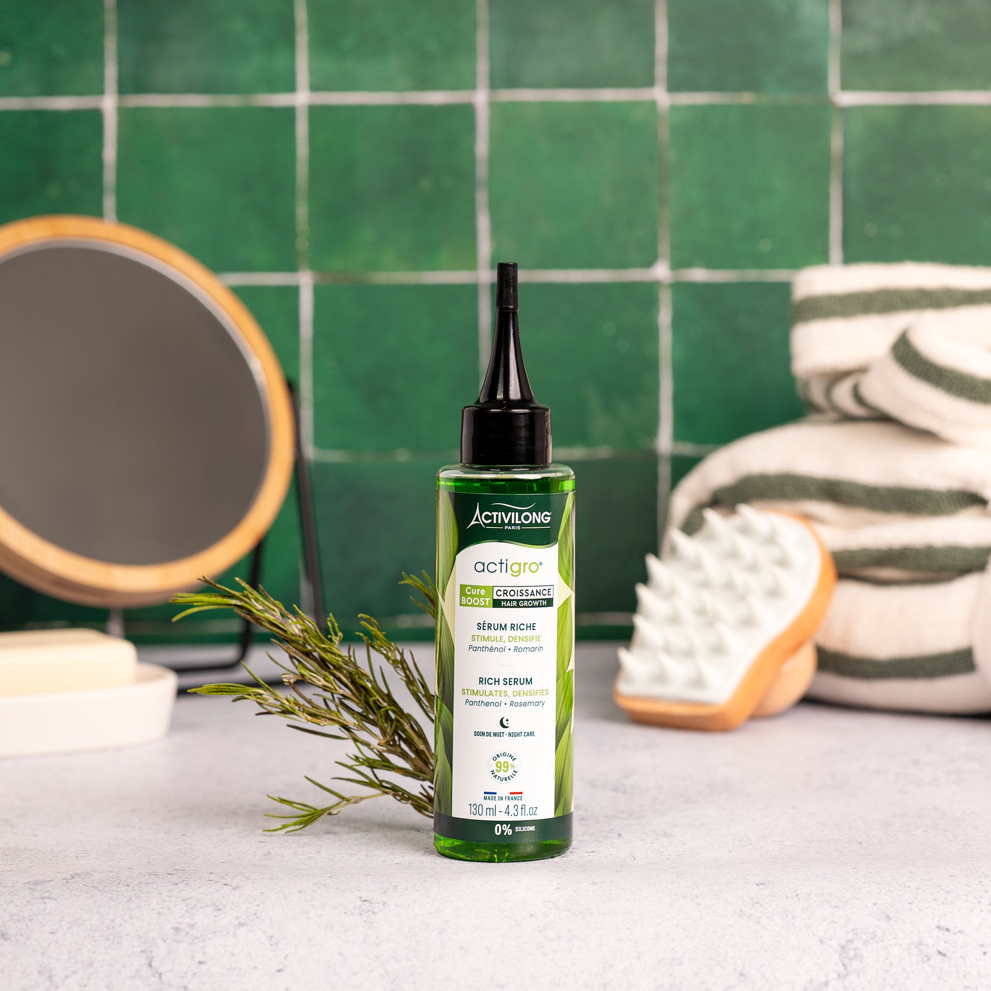 Visuel d'un Flacon de 130 ml du Sérum Riche Actigro sur un plan de salle de bain vert, avec miroir, brosse de massage et brin de romarin en arrière plan