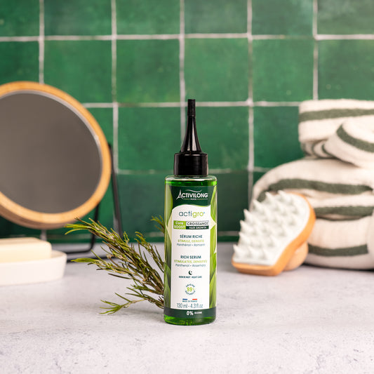Visuel d'un Flacon de 130 ml du Sérum Riche Actigro sur un plan de salle de bain vert, avec miroir, brosse de massage et brin de romarin en arrière plan