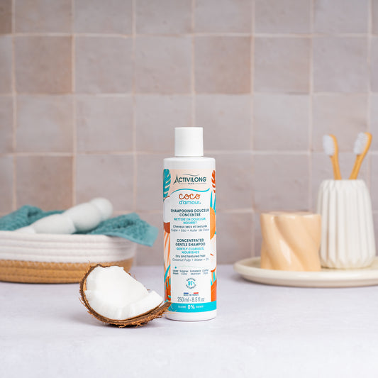 Flacon de Shampooing douceur concentré Coco d'Amour posé sur un plan de salle de bain, une demi-noix de coco fraîche à côté, avec un panier garni d'une serviette turquoise, un gobelet et des brosses à dents en arrière-plan, sur fond de carrelage beige.