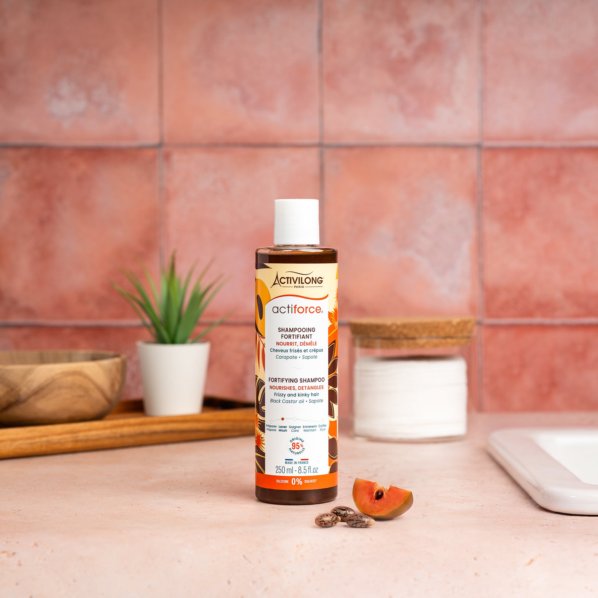 Visuel d'un Flacon 250 ml du Shampooing Fortifiant Actiforce sur un plan de salle de bain terracotta, avec graines de carapate et tranche de sapote en arrière plan