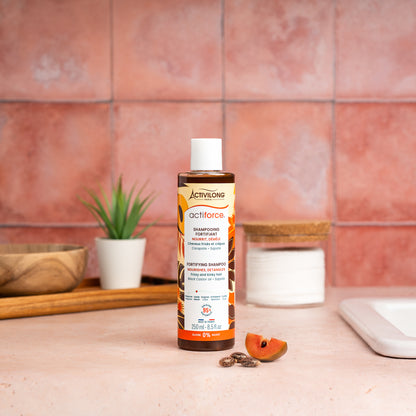 Visuel d'un Flacon 250 ml du Shampooing Fortifiant Actiforce sur un plan de salle de bain terracotta, avec graines de carapate et tranche de sapote en arrière plan
