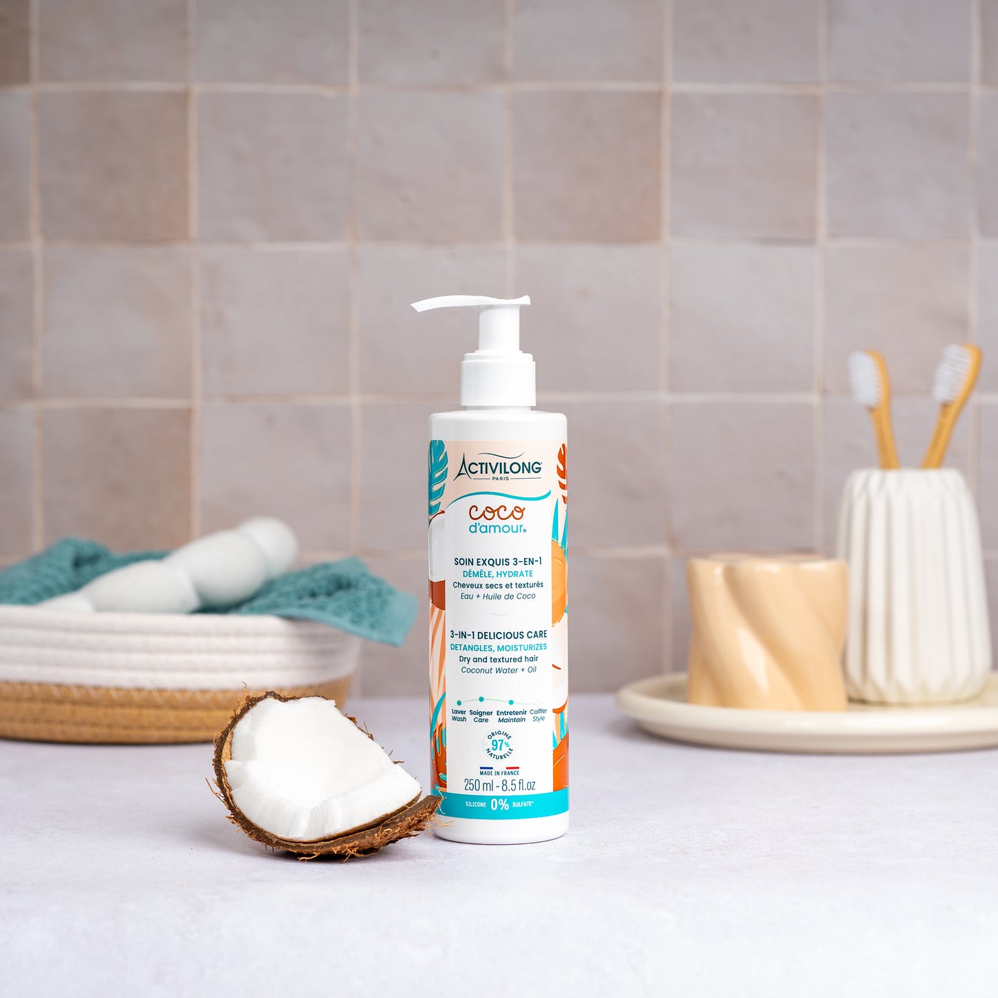 Flacon-pompe de Soin exquis 3-en-1 Coco d'Amour posé sur un plan de salle de bain, une demi-noix de coco fraîche à côté, avec un panier garni d'une serviette turquoise, un gobelet et des brosses à dents en arrière-plan, sur fond de carrelage beige.