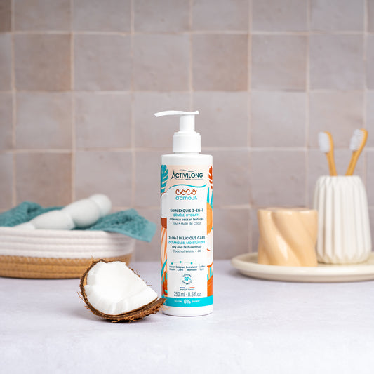 Flacon-pompe de Soin exquis 3-en-1 Coco d'Amour posé sur un plan de salle de bain, une demi-noix de coco fraîche à côté, avec un panier garni d'une serviette turquoise, un gobelet et des brosses à dents en arrière-plan, sur fond de carrelage beige.