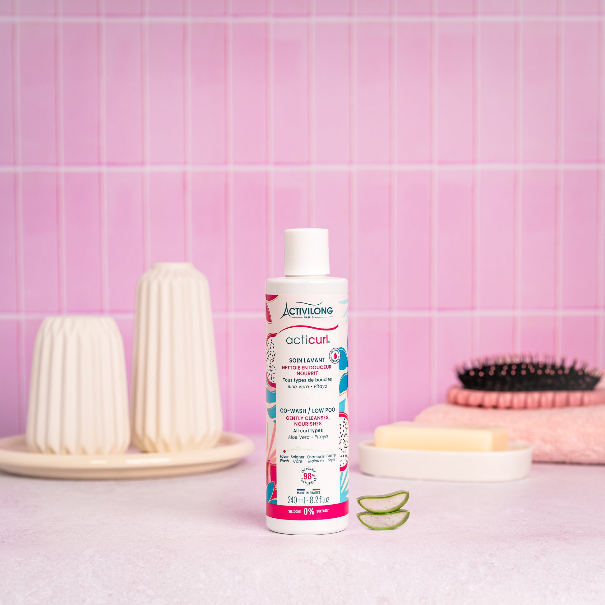 Visuel d'un Flacon du Soin Lavant / Co-Wash Acticurl sur un plan de salle de bain rose, avec brosse, savon et tranches d'aloe vera en arrière plan