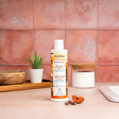 Visuel d'un Flacon de 240 ml du Soin Lavant / Co-Wash Actiforce sur un plan de salle de bain terracotta, avec graines de carapate et tranche de sapote en arrière plan